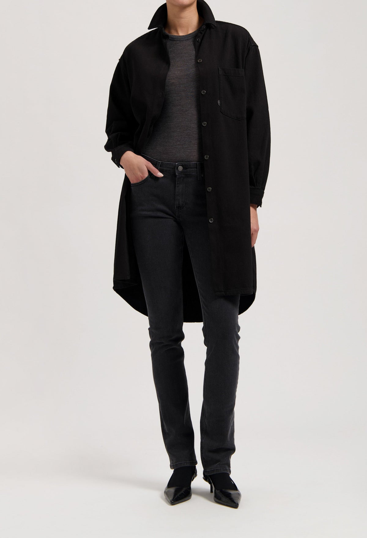 Model wearing a long black button-up coat over a gray top, paired with fitted black jeans and pointed black shoes.
