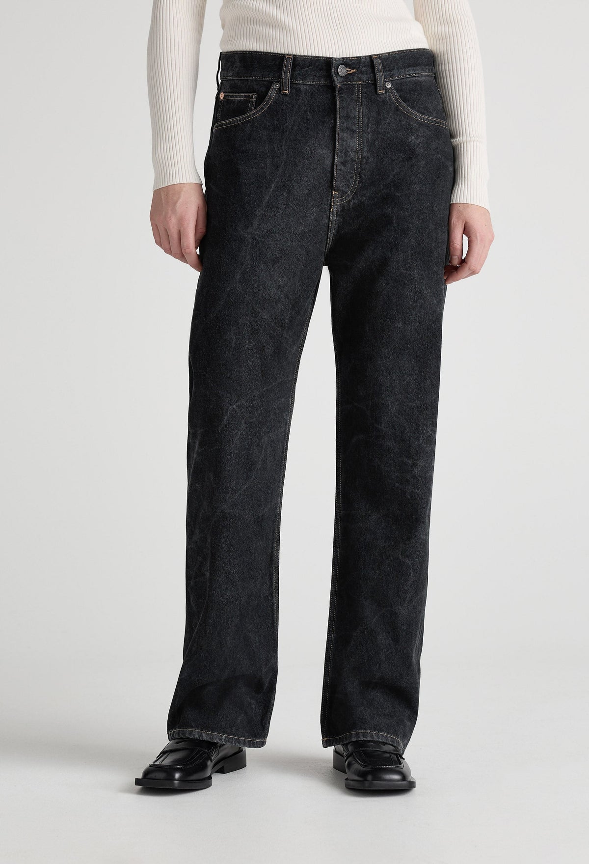 A model wearing loose-fitting black denim jeans with a ribbed, white long-sleeve top and black leather shoes.