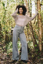 Woman wearing gray wide-leg denim pants and a beige knit sweater, standing in a sunlit forest trail.