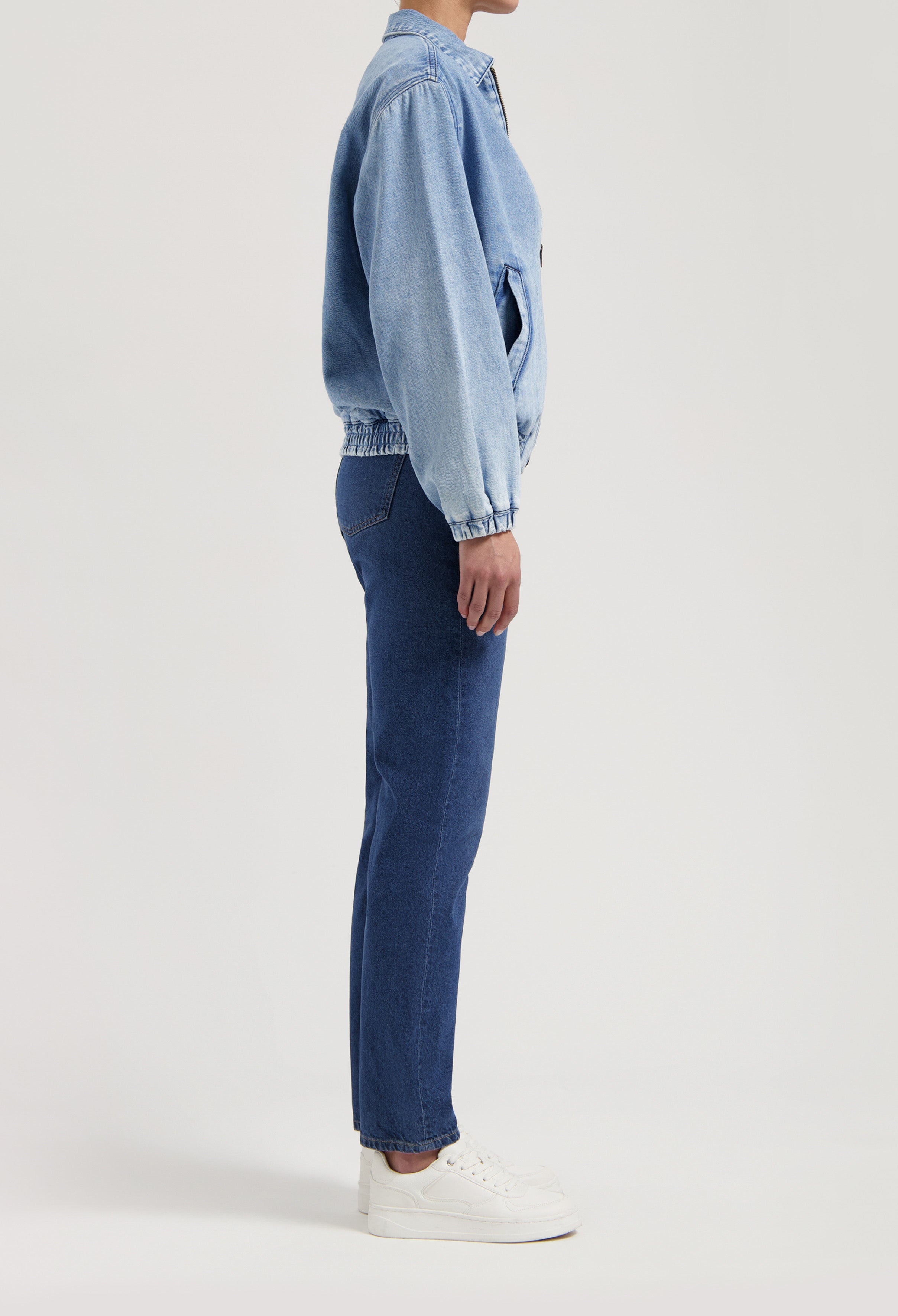 Side profile of a person in a light blue denim jacket and blue straight-leg jeans with white sneakers on a plain background.