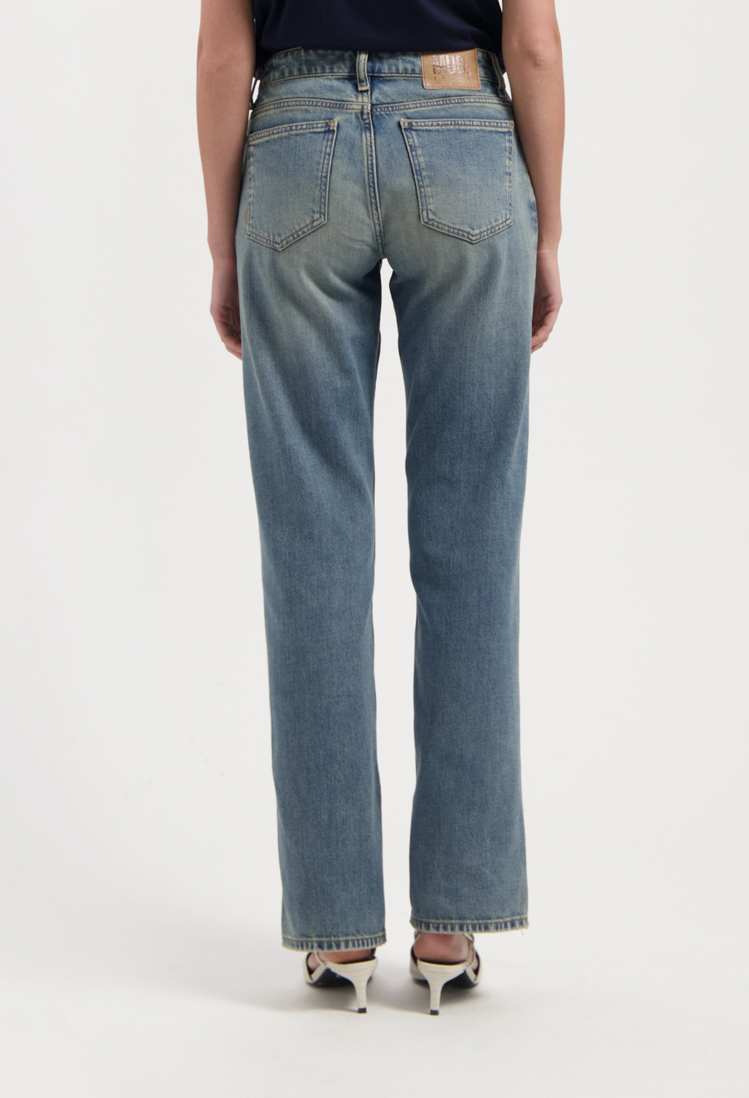 Back view of classic light blue high-waisted jeans paired with white open-back heels on a neutral background.