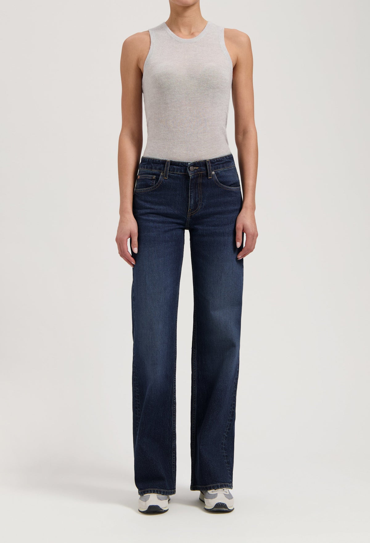 Woman wearing a light gray sleeveless top paired with dark blue, wide-leg jeans and white sneakers, standing in studio.