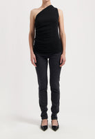 Model in sleek black one-shoulder top and form-fitting dark jeans, featuring pointed black heels on a minimalist backdrop.