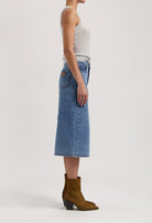 Side view of a woman in a sleeveless gray top, wearing a blue denim midi skirt paired with brown suede ankle boots.