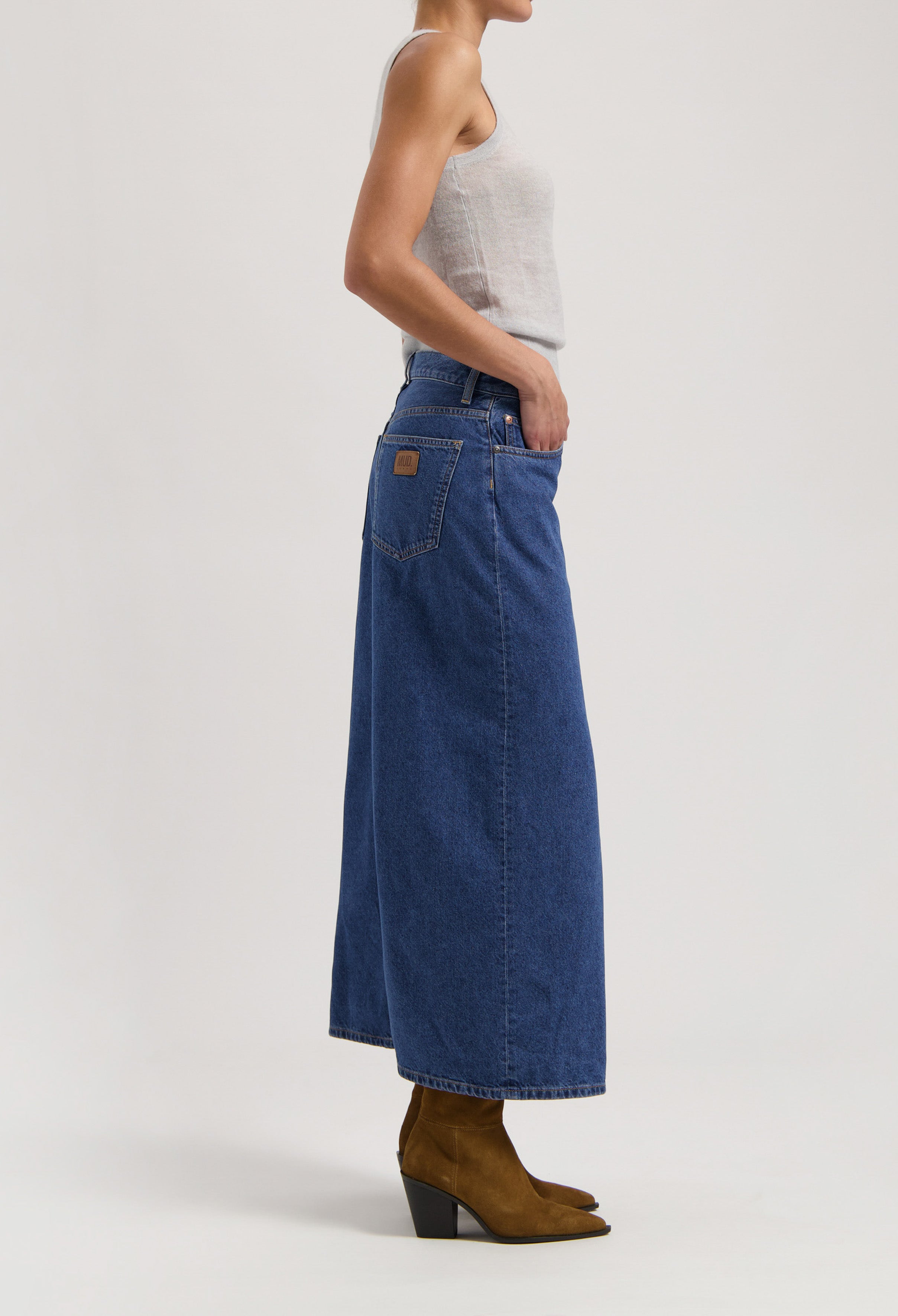 Woman wearing a sleeveless gray top and high-waisted blue denim maxi skirt paired with brown leather ankle boots.